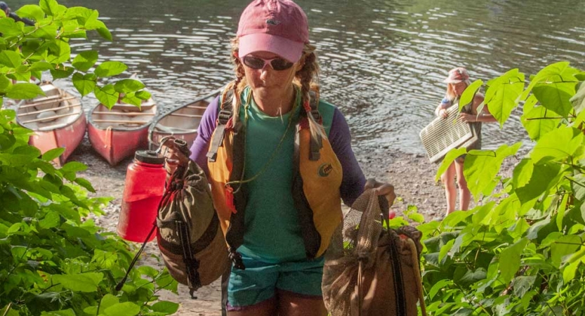 A person carries gear away from a shore. There are beached canoes in the background. 